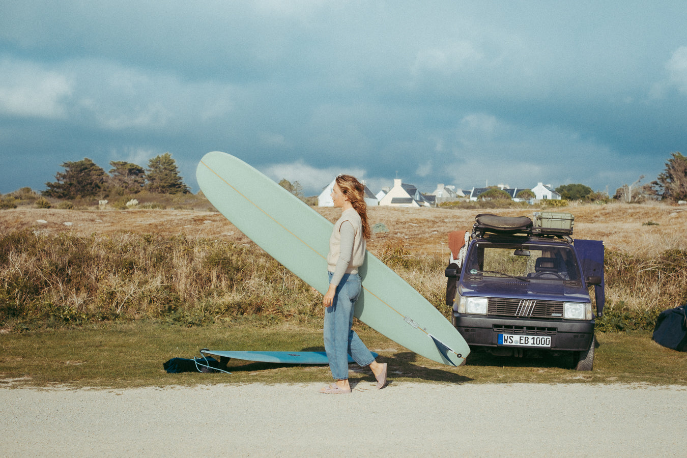 A woman holding a surf board walks past a Fiat Panda converted into a van