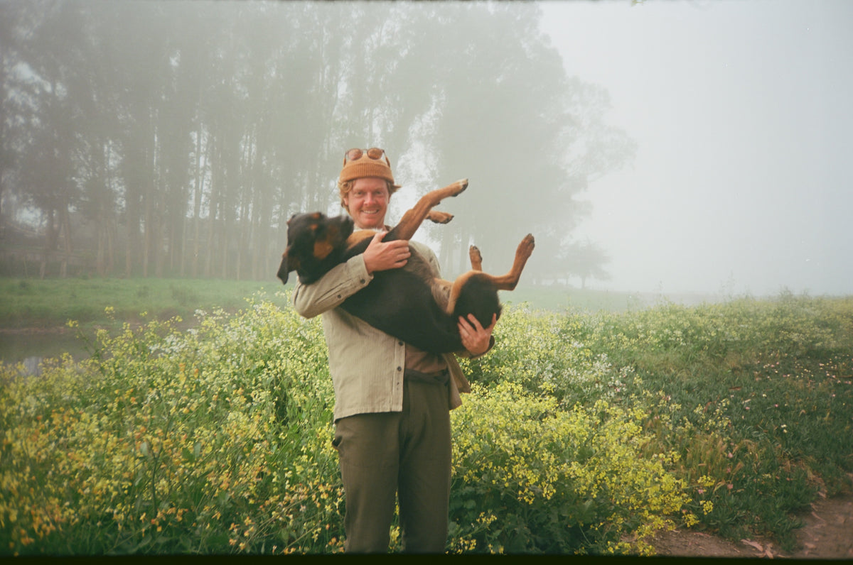 A man wearing a tan hat and light-colored shirt holds a black and tan dog upside down in a misty outdoor field with yellow flowers and trees in the background.