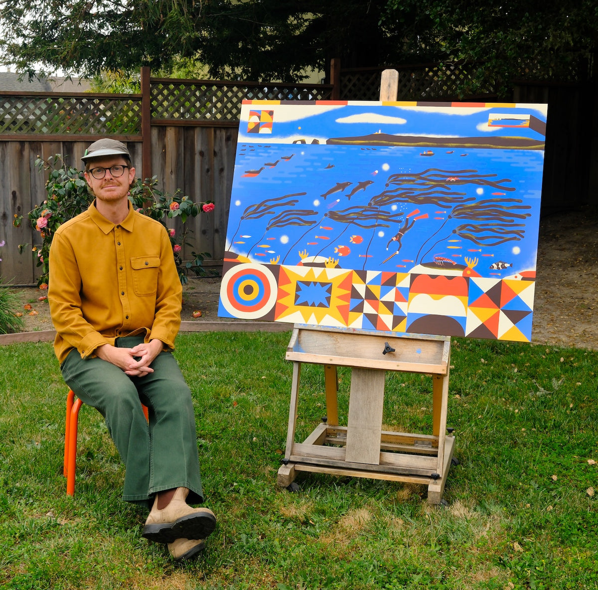 A man wearing a mustard yellow shirt, green pants, and a gray cap sits on an orange chair next to a large colorful painting on a wooden easel in a backyard setting.