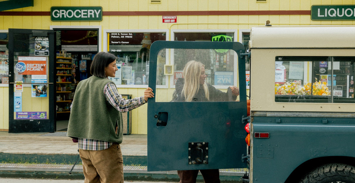 Two women looking in the back of a truck, parked outside a grocery store