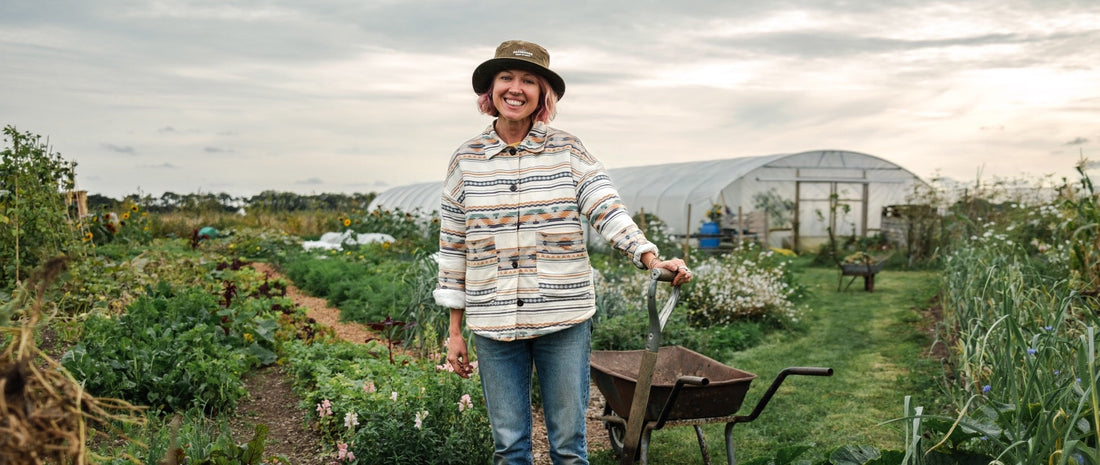 A woman stands with a spade by a wheelbarrow  surrounded by rows of vegetables and plants