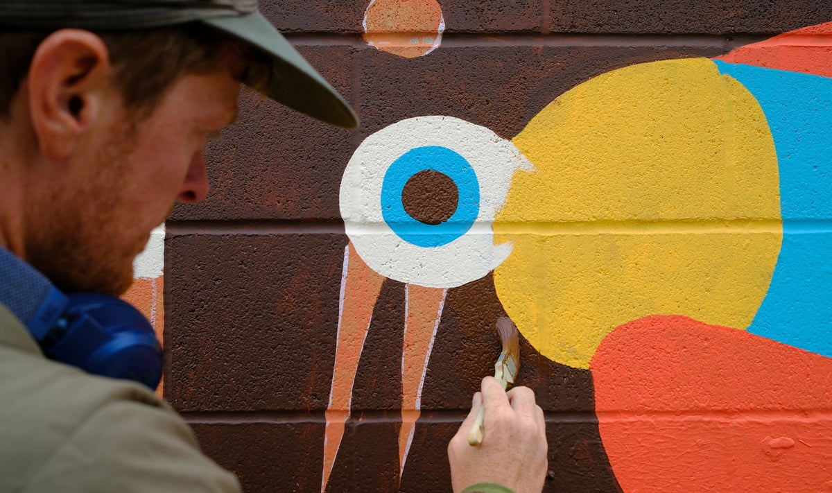 A man paints a colourful bird mural on a brick wall