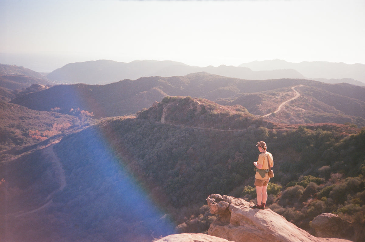 A person stands on a rocky cliff overlooking a vast mountain landscape with winding roads and hazy hills under a bright sky.
