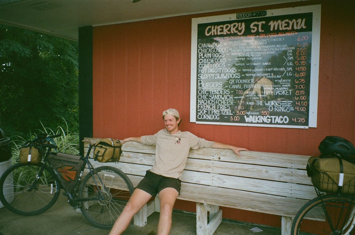 A man sits on a wooden bench in front of a red wall with a large 'Cherry St. Menu' chalkboard sign, with two bicycles on either side of him.