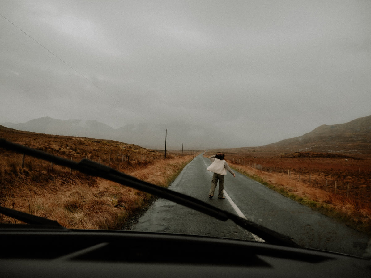 A person stands on a wet country road between hills under an overcast sky, viewed from inside a vehicle with windshield wipers visible in the foreground.