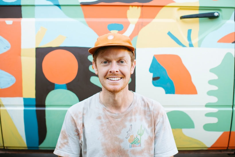 A smiling man wearing a light gray tie-dye t-shirt and an orange cap stands in front of a colorful abstract mural painted on a vehicle.